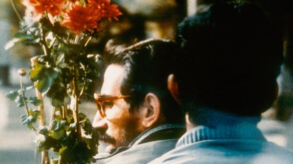 Close-Up film image; two men standing close to each other with a bouquet of flowers in front of them