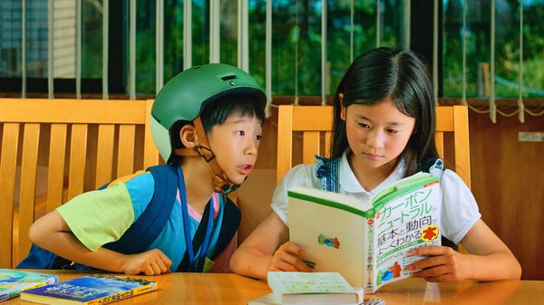 How Dare You? film image; young boy in a helmet leaning into a girl reading a book