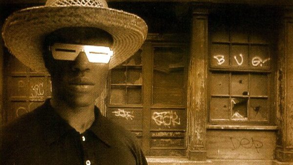 sepia-toned image of man in a hat and funky glasses in front of a dilapidated building