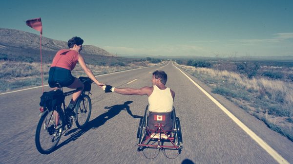Man in Motion: The Rick Hansen Story film image; man in a wheelchair reaching out to man on bike