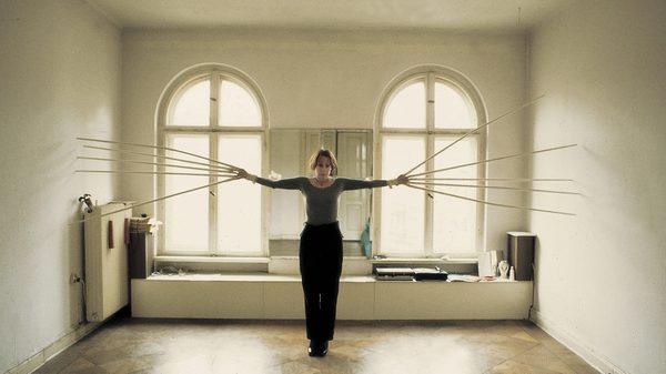 Rebecca Horn film image; woman in a empty room holding out her arms with makeshift scissorhands
